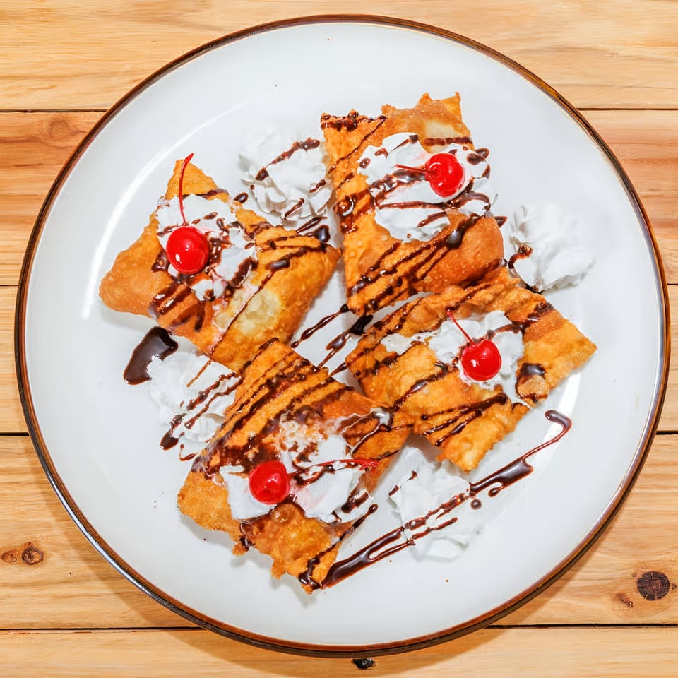 Four golden-fried dessert chimichangas topped with whipped cream, chocolate drizzle, and a cherry on a white plate.