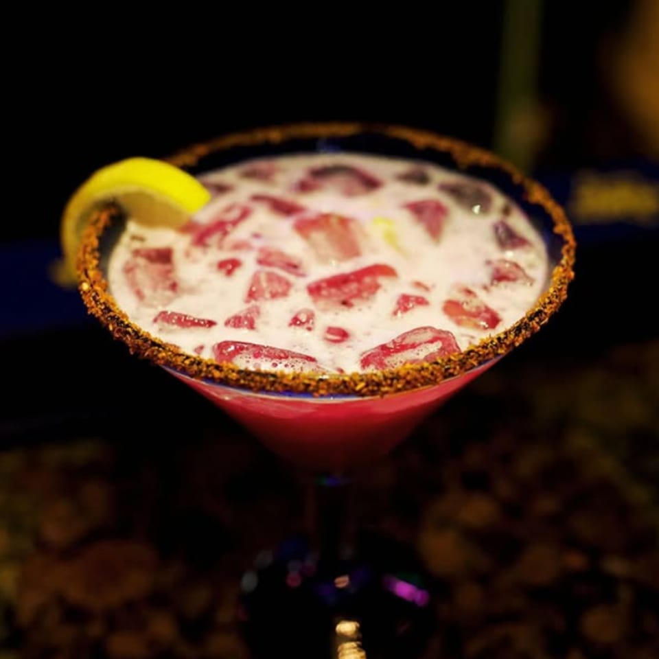 Red cocktail with ice, foam, a lemon slice, and a chili-salted rim in a martini glass.