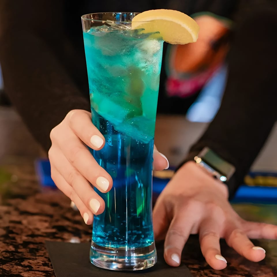 A bartender's hands serving a tall glass of vibrant blue cocktail with ice and a lemon wedge.