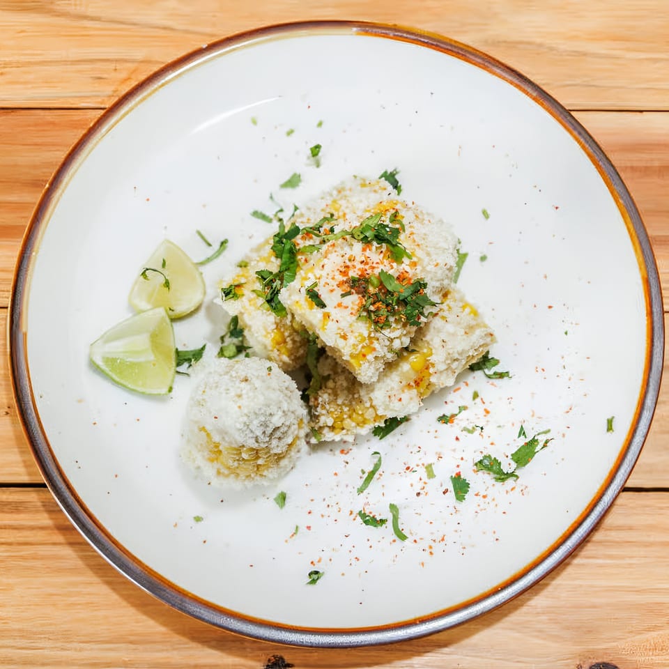 Elote, Mexican street corn, on a white plate with lime wedges, cilantro, and chili powder.