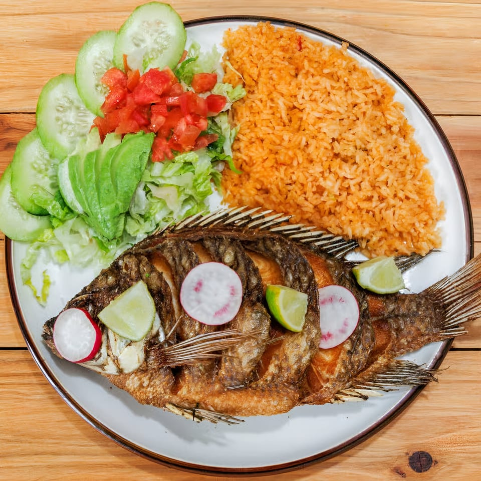 Fried whole fish with rice, salad, avocado, radishes, and lime on a white plate.