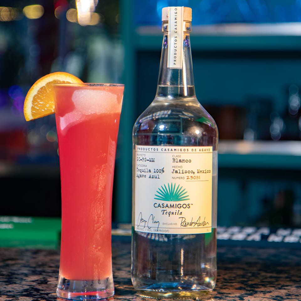 A tall red cocktail with an orange slice next to a bottle of Casamigos Blanco Tequila.